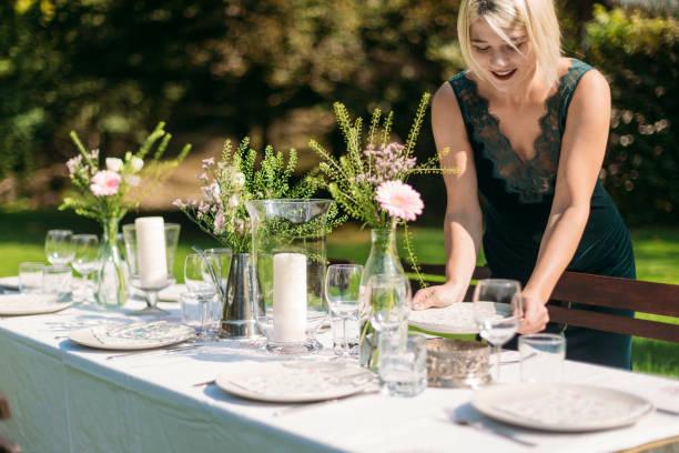 Young blonde woman setting and decorating table for lunch outdoors on a sunny day.