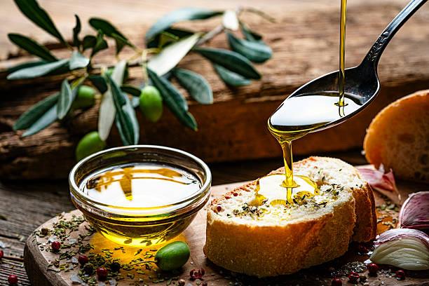 Mediterranean food: Extra virgin olive oil pouring from spoon to a bread slice on rustic wooden table. Green olives, pepper, salt and garlic. Savory food