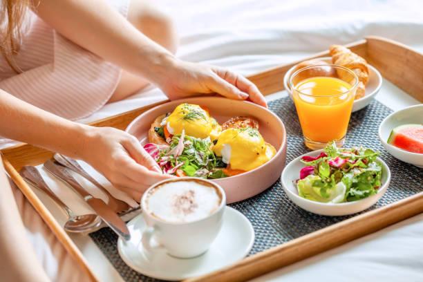 Breakfast in Bed Served with Cup of Coffee, Salad, Fresh Fruits and Eggs Benedict on Wooden Tray. Woman Hands Holding Plate with Fresh Food. Room Service in Hotel